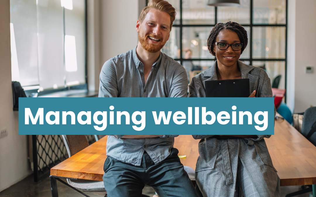 Young man and young woman sit on edge of table in workplace. They are both smiling and looking directly into the camera. Text reads managing wellbeing.